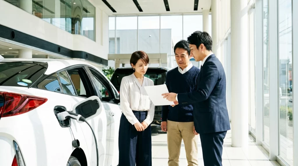 ショールームで、日本人夫婦がハイブリッド車の横で販売員から見積書を受け取っている